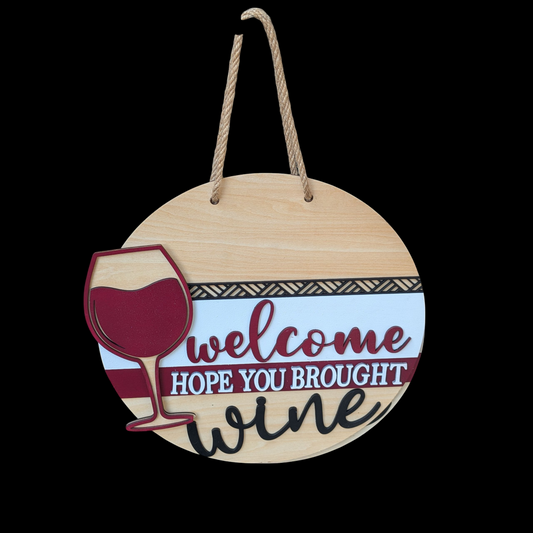 Wooden round sign with a wine glass graphic and text 'Welcome, hope you brought wine' on a natural wood background.
